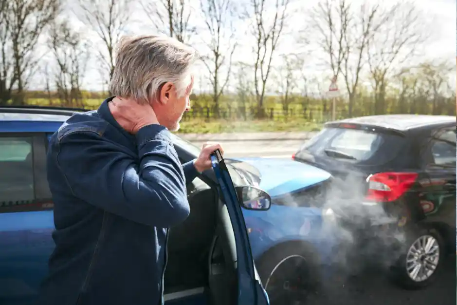 An older man stands beside a car after a collision, holding his neck in discomfort. The car he was in has been rear-ended, with smoke rising from the engine.