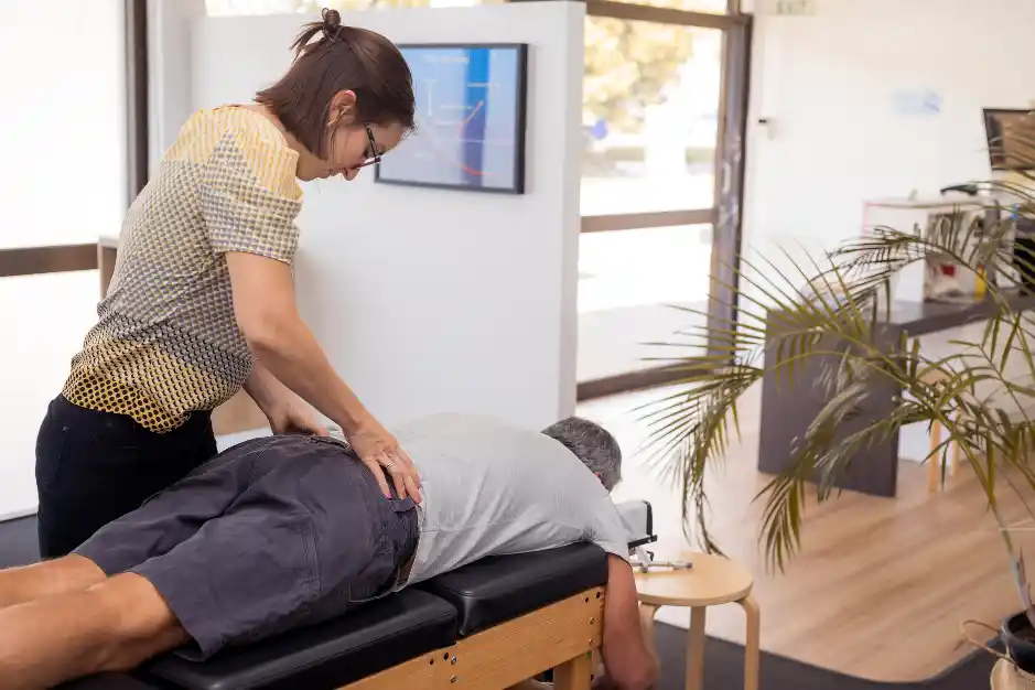 A chiropractor is performing an adjustment on a patient lying face down on a treatment table. The patient’s lower back is being gently manipulated, and the setting is a bright, professional clinic.