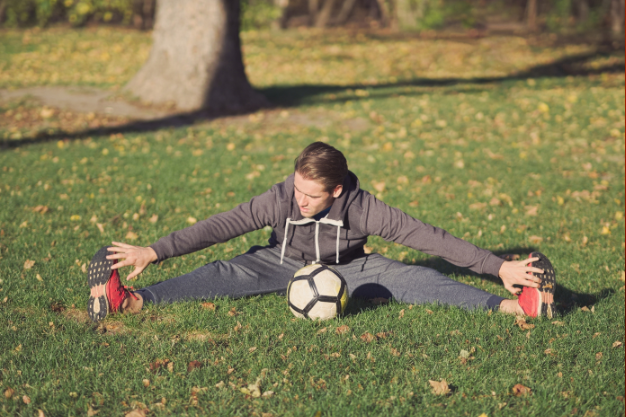 A person is stretching their legs wide apart on a grassy field, preparing for a soccer game. A soccer ball is positioned in front of them as they reach forward.