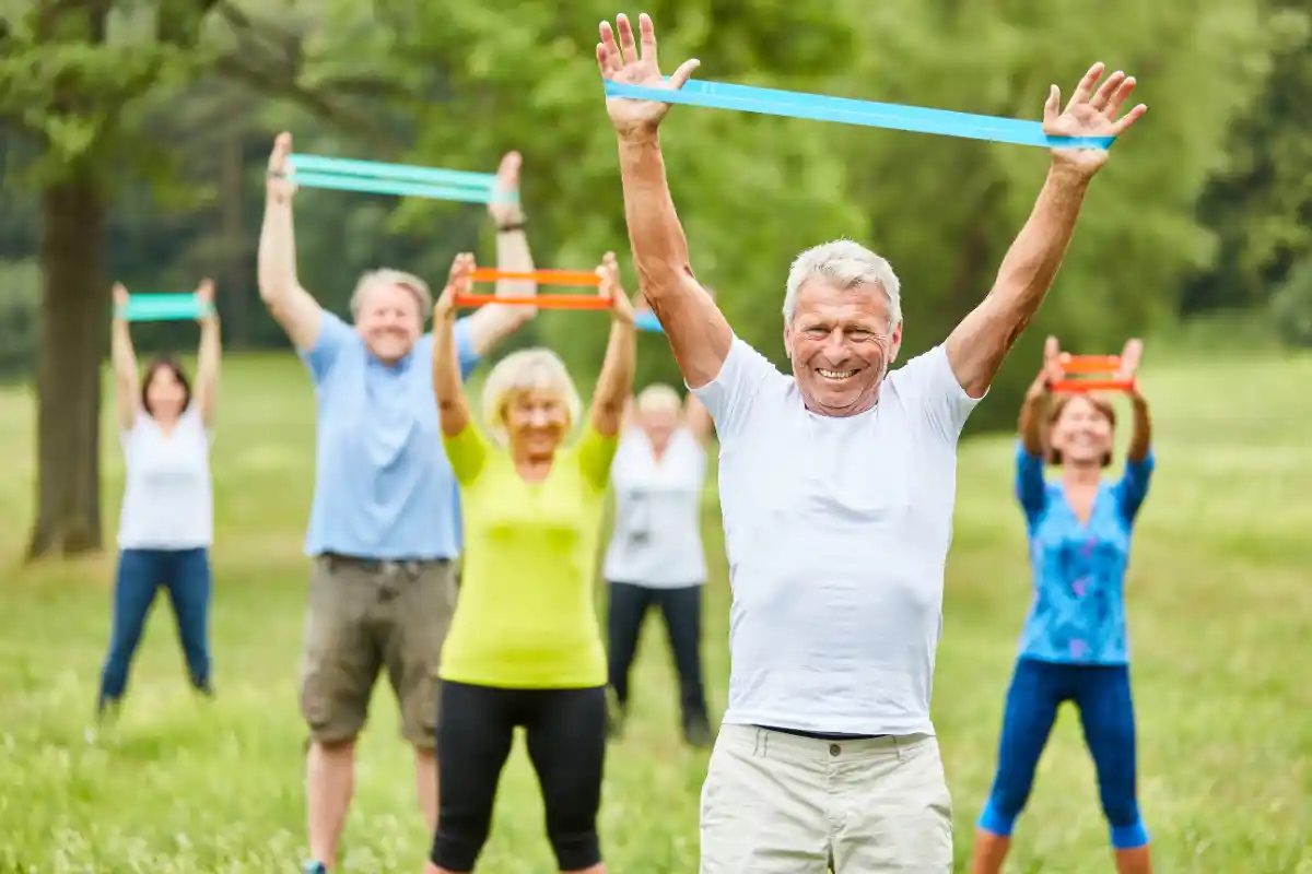 A group of older adults are exercising outdoors, using resistance bands. They are stretching and lifting their arms while smiling, in a lush green park setting.
