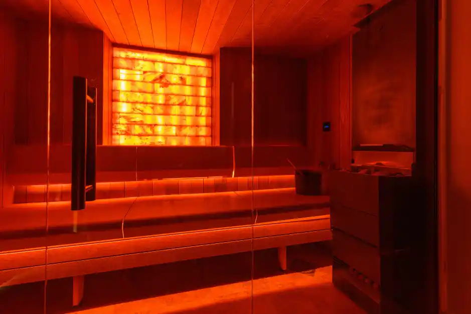 A warm and relaxing sauna with orange lighting. The room features a wooden bench, a stone wall, and a stove, creating a tranquil environment.