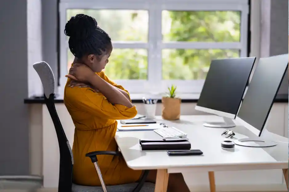 A woman sitting at a desk with two monitors rubs her neck in discomfort, possibly from sitting in the same position for too long. The workspace looks tidy, with a view of greenery outside.