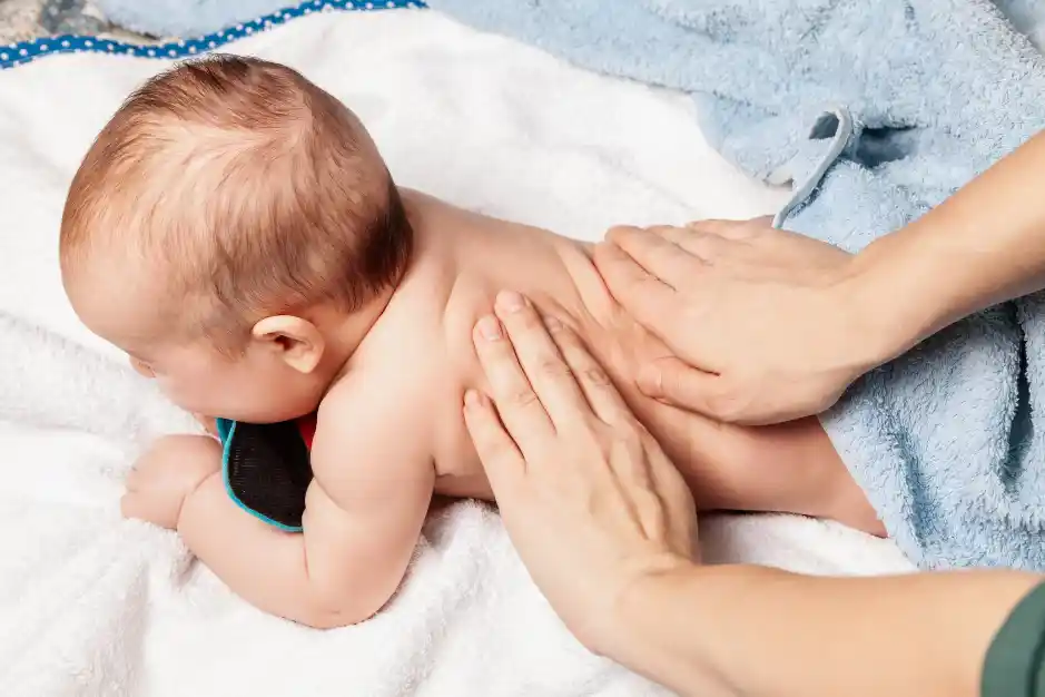 A baby is receiving a gentle back massage, with a caregiver's hands supporting and soothing the baby. The setting is calm with soft towels.